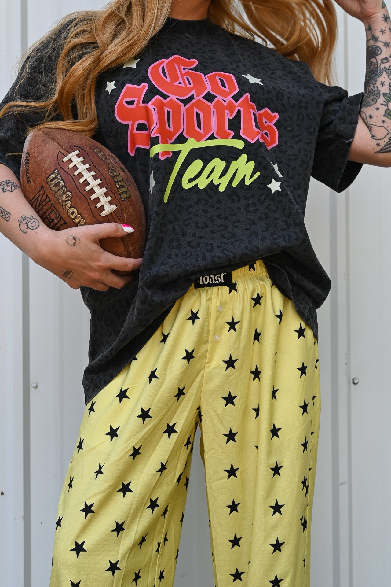 A person wearing a black graphic T-shirt that reads 'Go Sports Team' in red, white, and green, paired with yellow pants covered in black stars. The person is holding a brown Wilson football and standing against a light gray metal wall