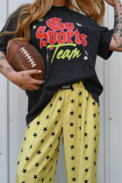 A person wearing a black graphic T-shirt that reads 'Go Sports Team' in red, white, and green, paired with yellow pants covered in black stars. The person is holding a brown Wilson football and standing against a light gray metal wall