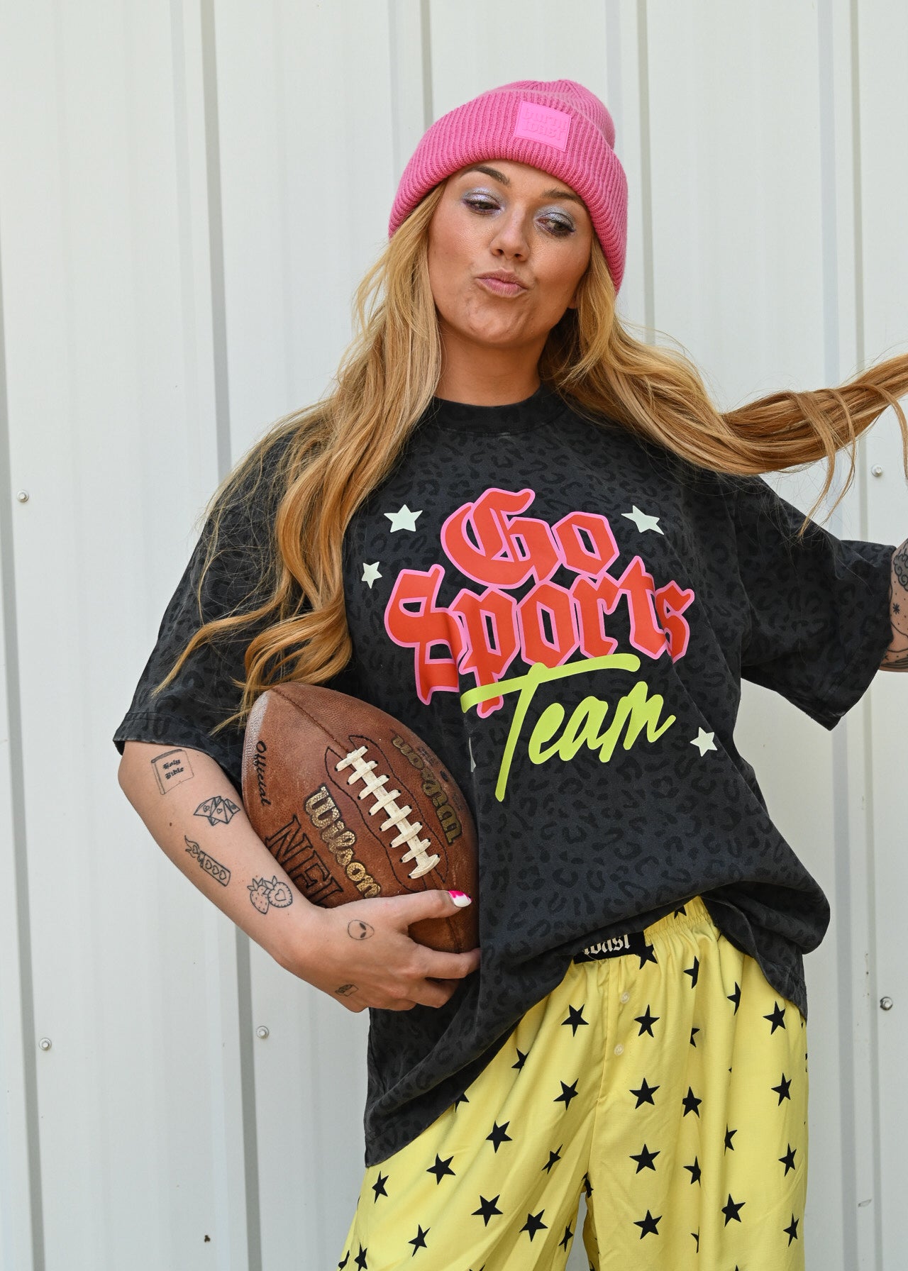 A woman wearing a pink beanie, a black graphic t-shirt that says 'Go Sports Team' in red and green, and yellow pants with black stars, holding a football against a white corrugated metal wall