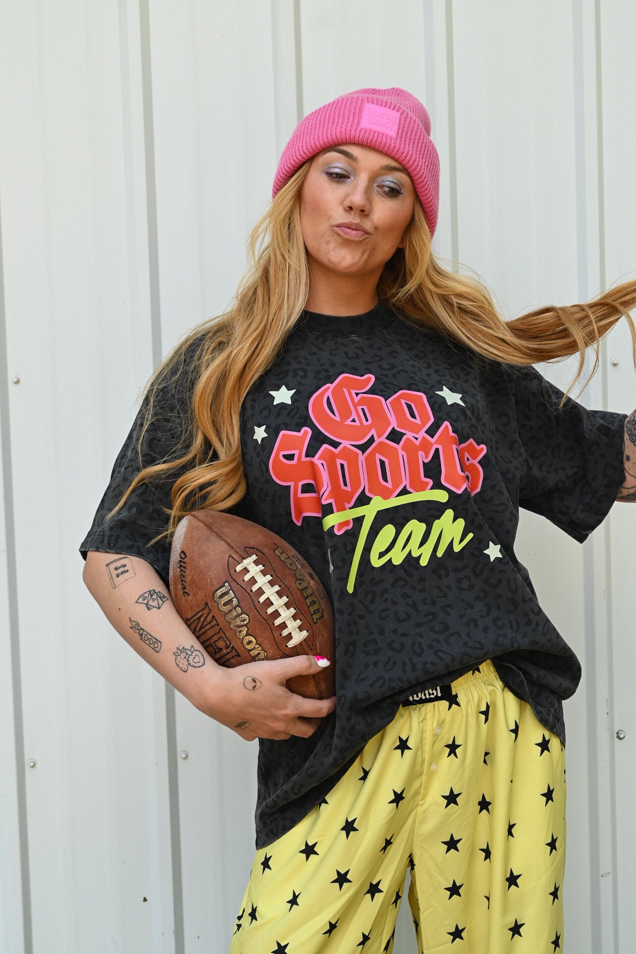 A woman wearing a pink beanie, a black graphic t-shirt that says 'Go Sports Team' in red and green, and yellow pants with black stars, holding a football against a white corrugated metal wall