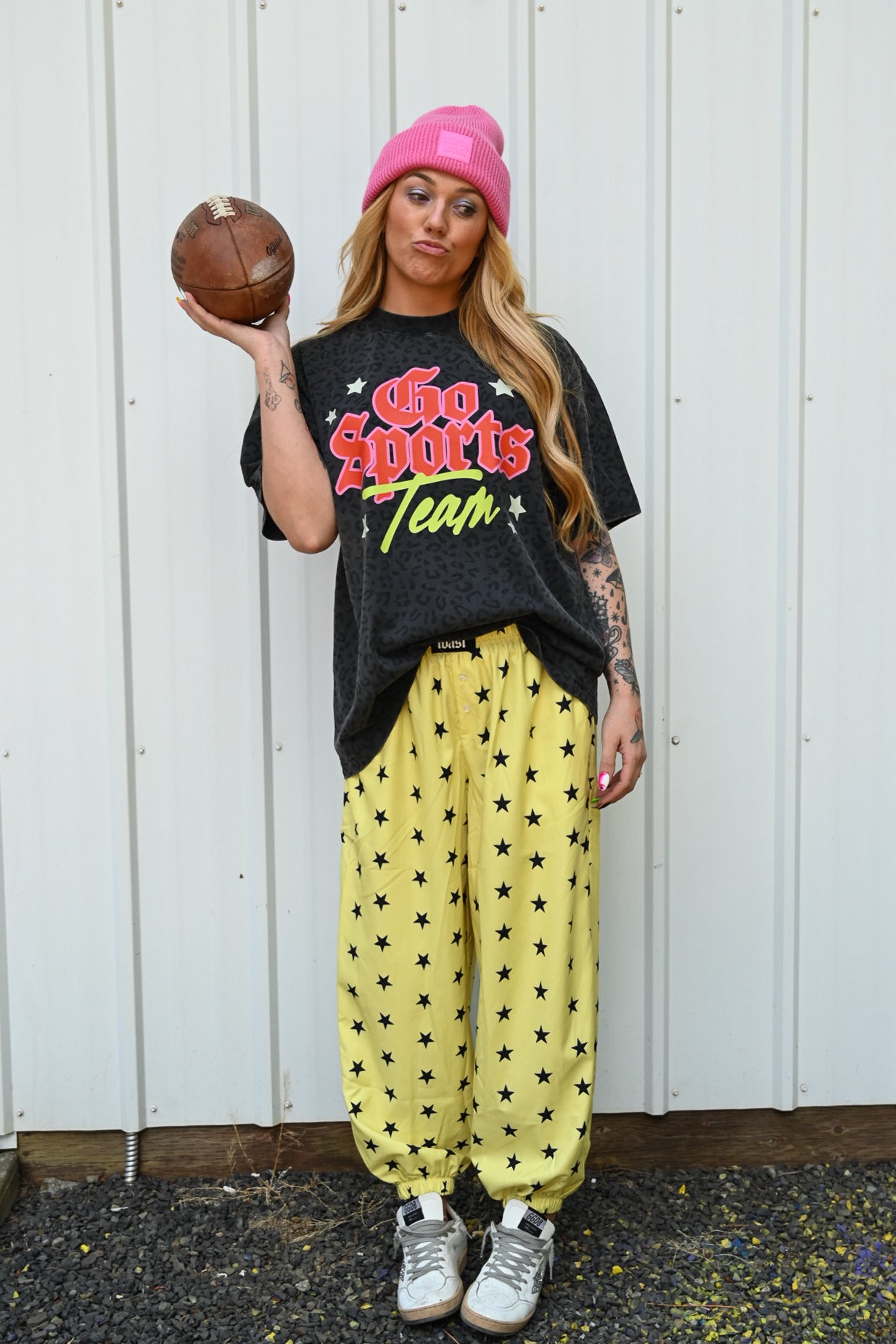 A woman stands in front of a white corrugated metal wall holding a brown football. She is wearing a pink beanie, a black graphic t-shirt that reads 'Who Sports Team', yellow sweatpants with black star patterns, and gray sneakers