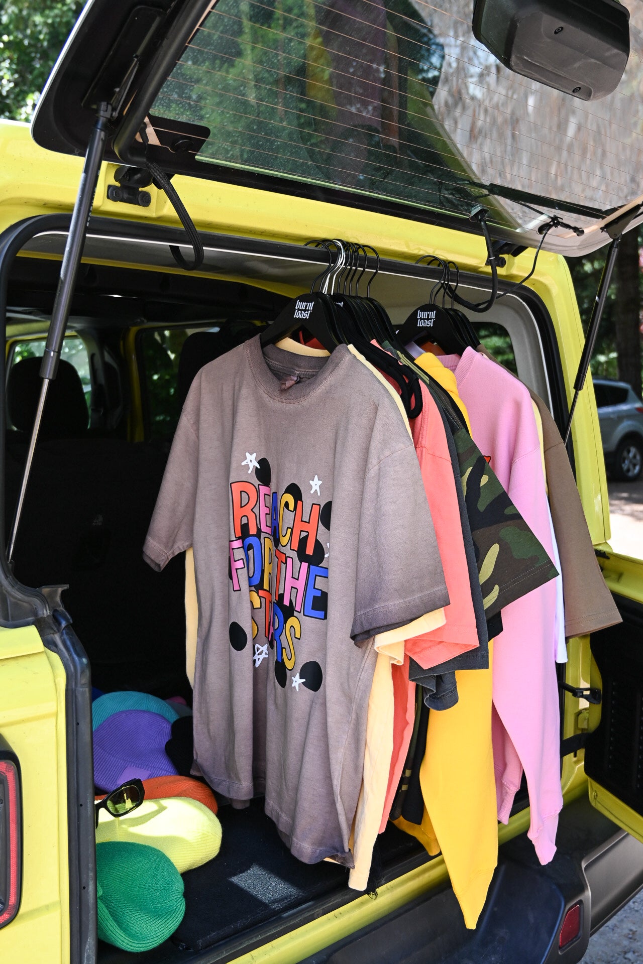 A gray oversized graphic T-shirt with colorful retro text hangs in the open trunk of a bright yellow vehicle, alongside other colorful T-shirts. Hats and sunglasses are stacked on the floor of the trunk
