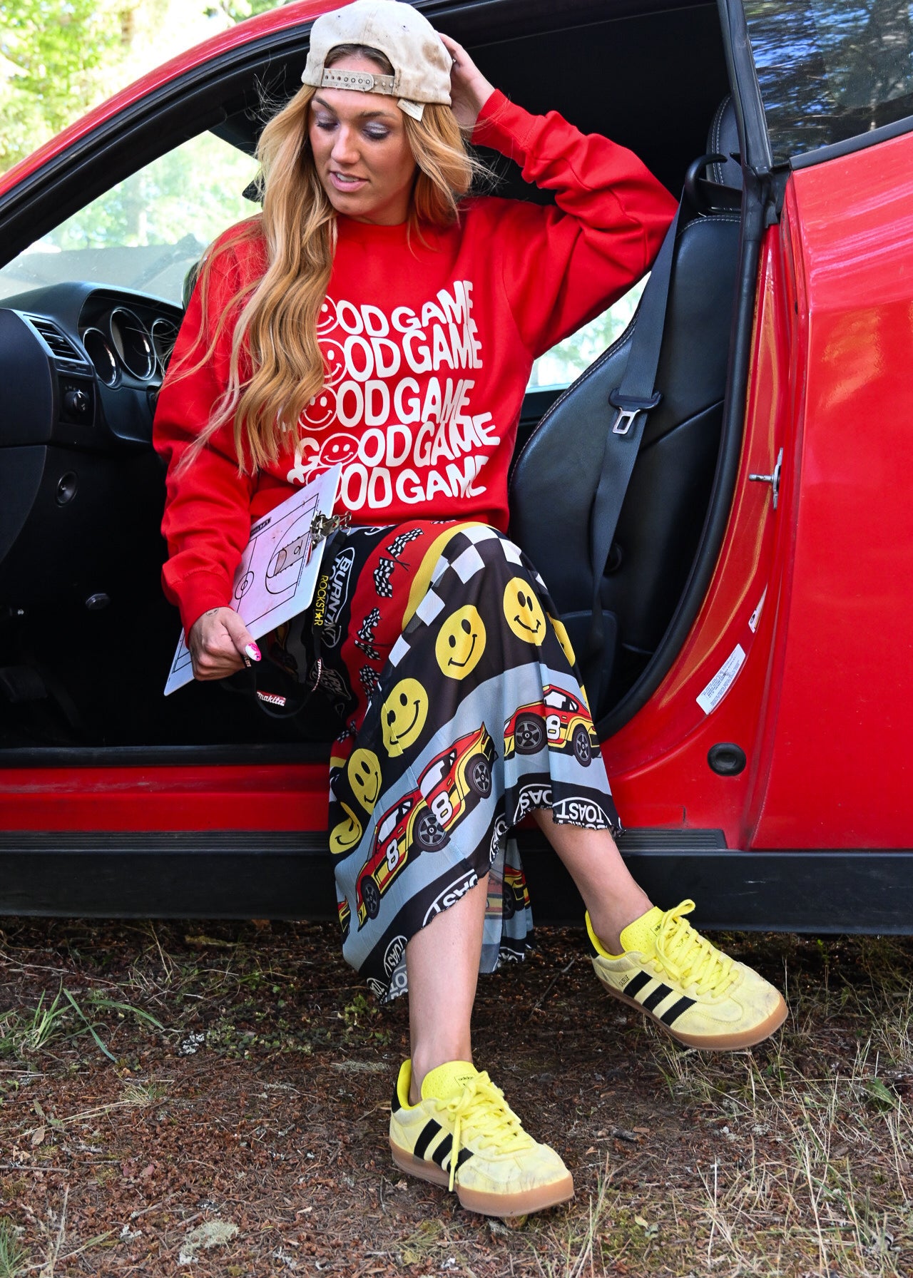 A woman sitting in the open door of a red car outdoors, wearing a red sweatshirt with white 'GOOD GAME' text, a patterned skirt, and yellow sneakers