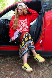 A woman sitting in the open door of a red car outdoors, wearing a red sweatshirt with white 'GOOD GAME' text, a patterned skirt, and yellow sneakers