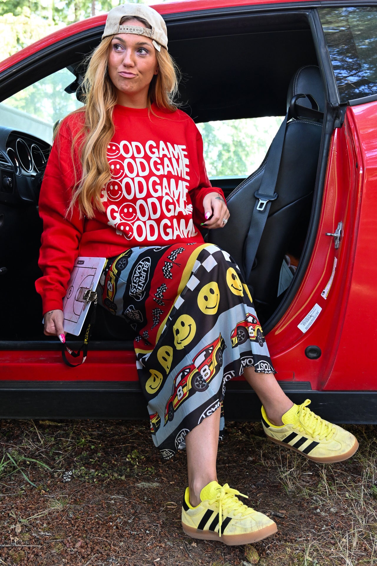 A woman sitting in the open door of a red car, wearing a red sweatshirt with white 'GOOD GAME' text, a patterned skirt, and yellow Adidas sneakers
