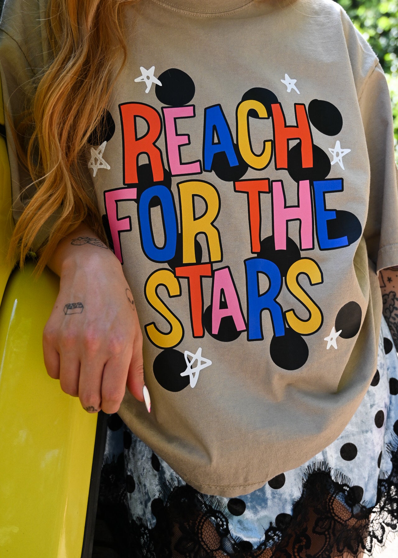A person wearing a beige oversized graphic T-shirt with the phrase “REACH FOR THE STARS” in large multicolored letters, surrounded by black polka dots and white stars. The photo is cropped to show the torso, hair, and part of a patterned skirt, with a bright yellow car visible on the left