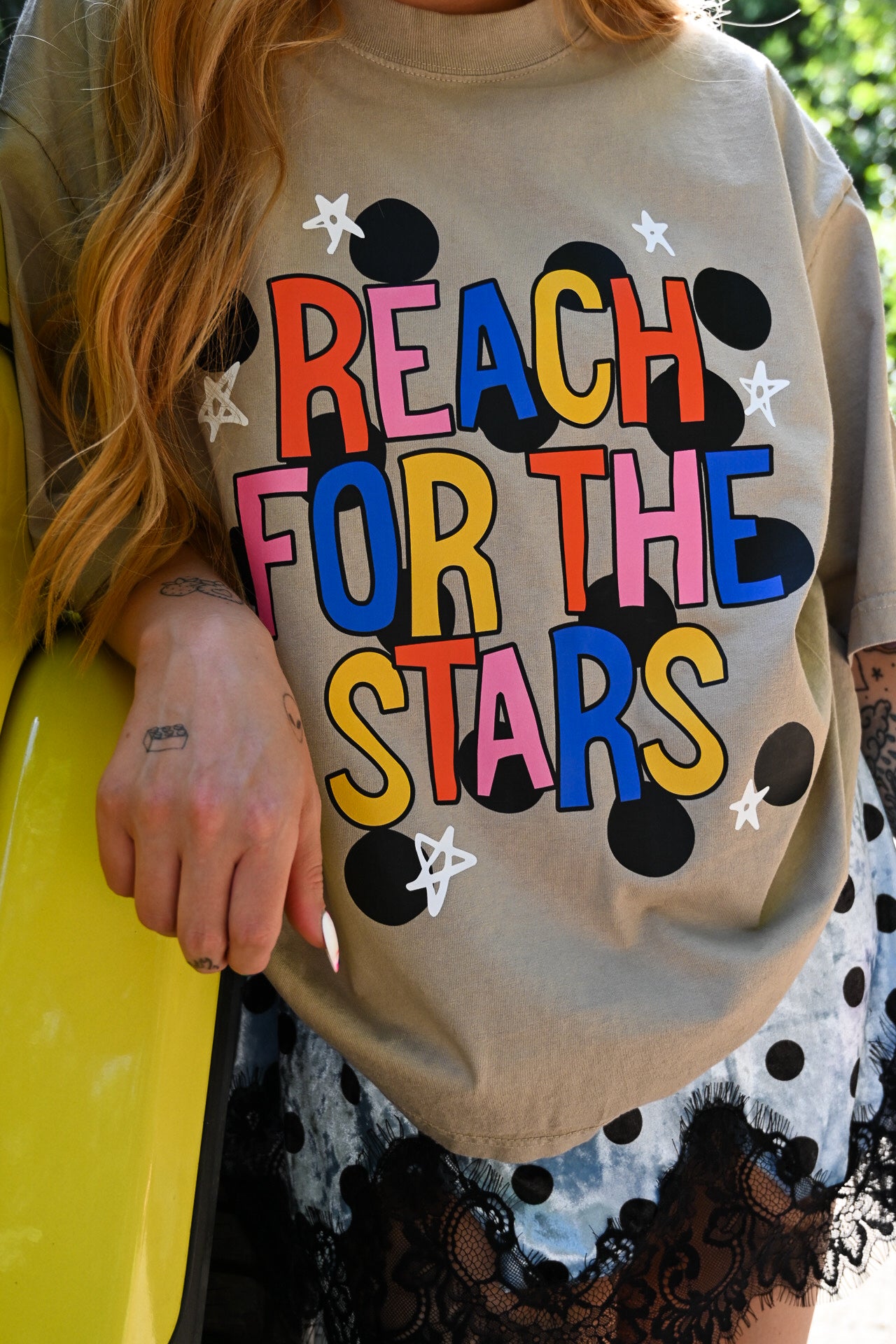 A person wearing a beige oversized graphic T-shirt with the phrase “REACH FOR THE STARS” in large multicolored letters, surrounded by black polka dots and white stars. The photo is cropped to show the torso, hair, and part of a patterned skirt, with a bright yellow car visible on the left