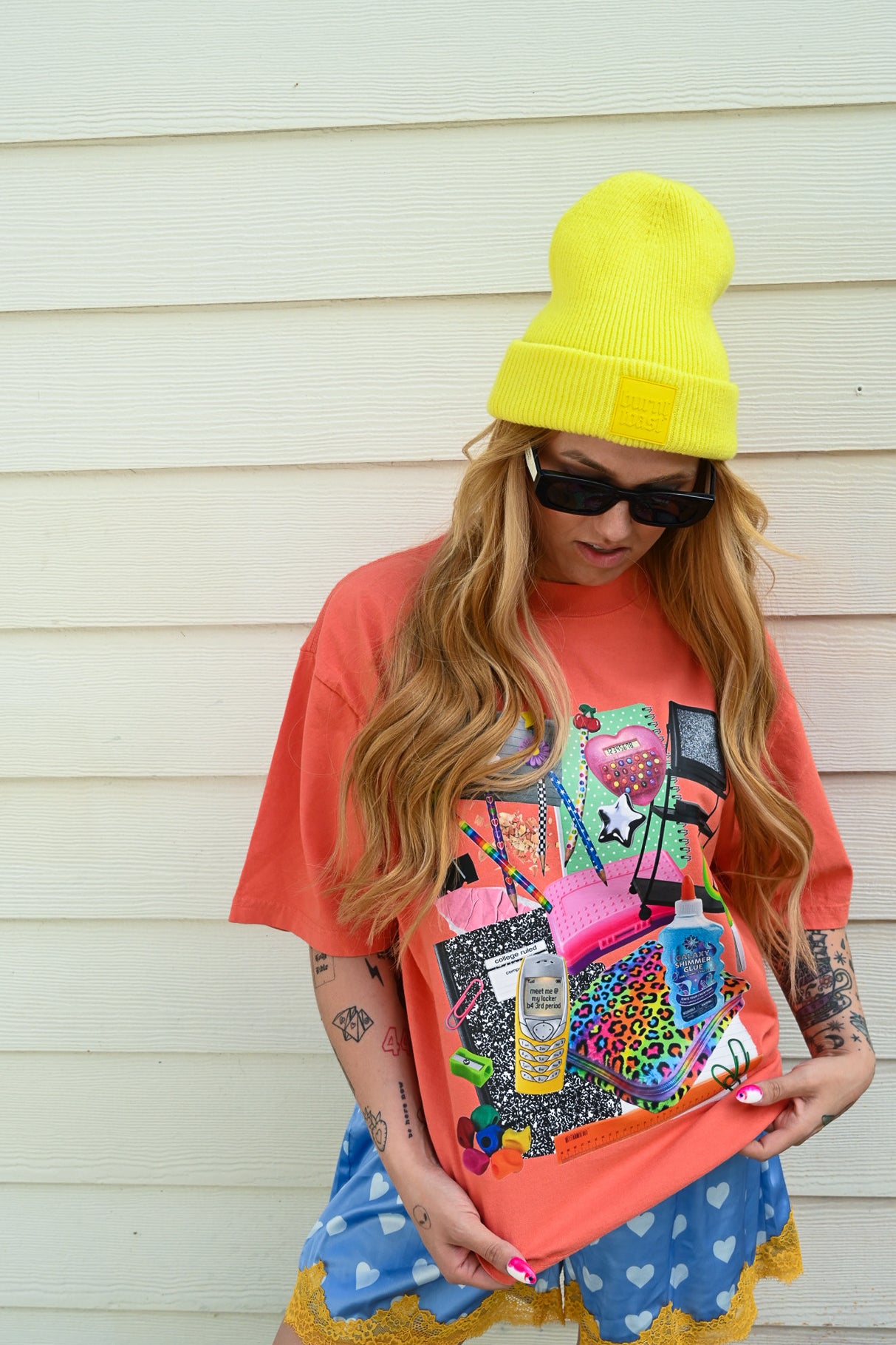 A woman wearing a bright yellow beanie, black sunglasses, and a coral orange graphic t-shirt holds up a matching coral orange pouch decorated with colorful pop culture stickers and graphics. She is standing against a white wood-paneled wall