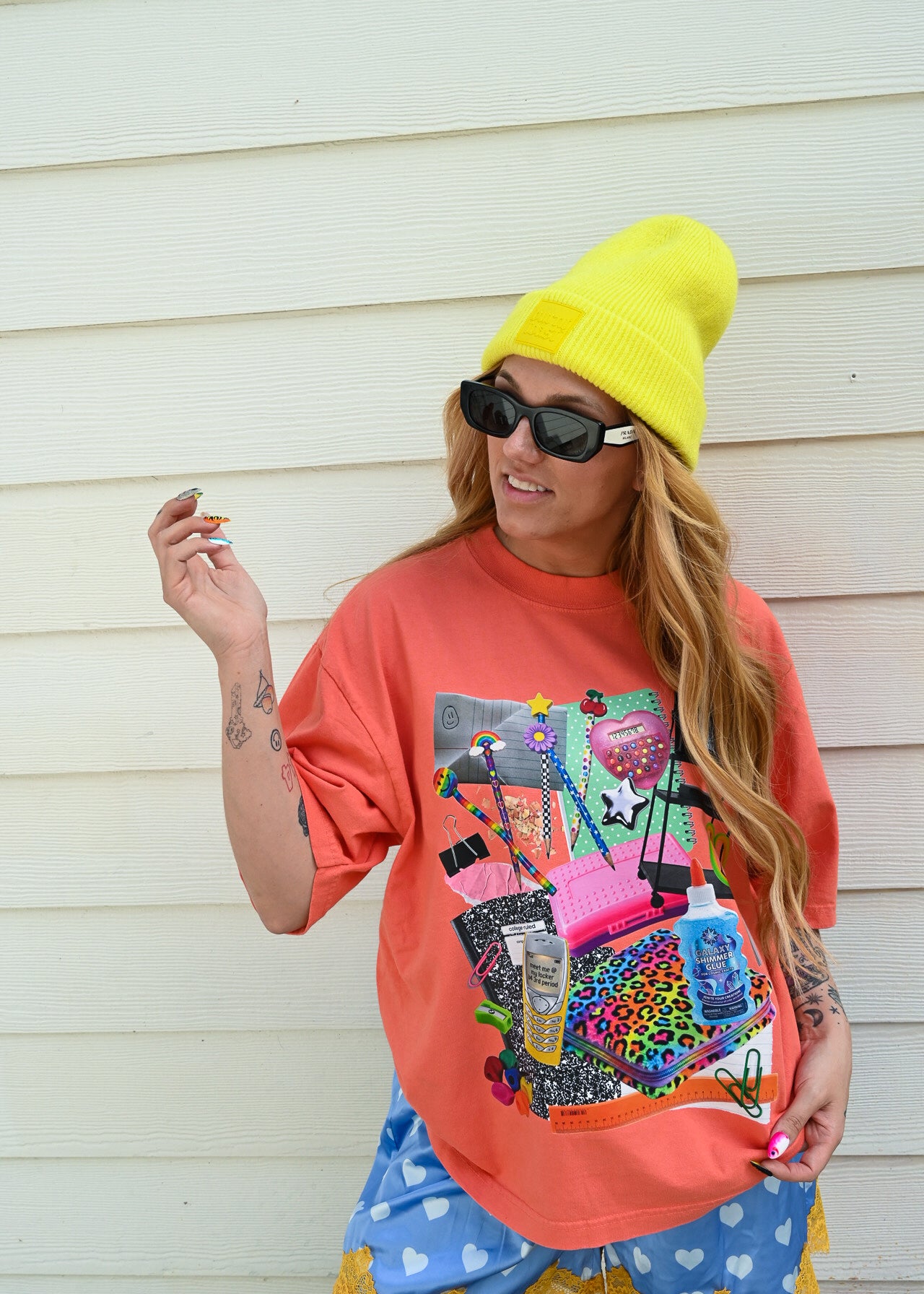 A woman wearing a bright yellow beanie, black sunglasses, a coral orange graphic t-shirt, and blue polka-dot shorts with yellow ruffle trim, standing against a white wood-paneled wall