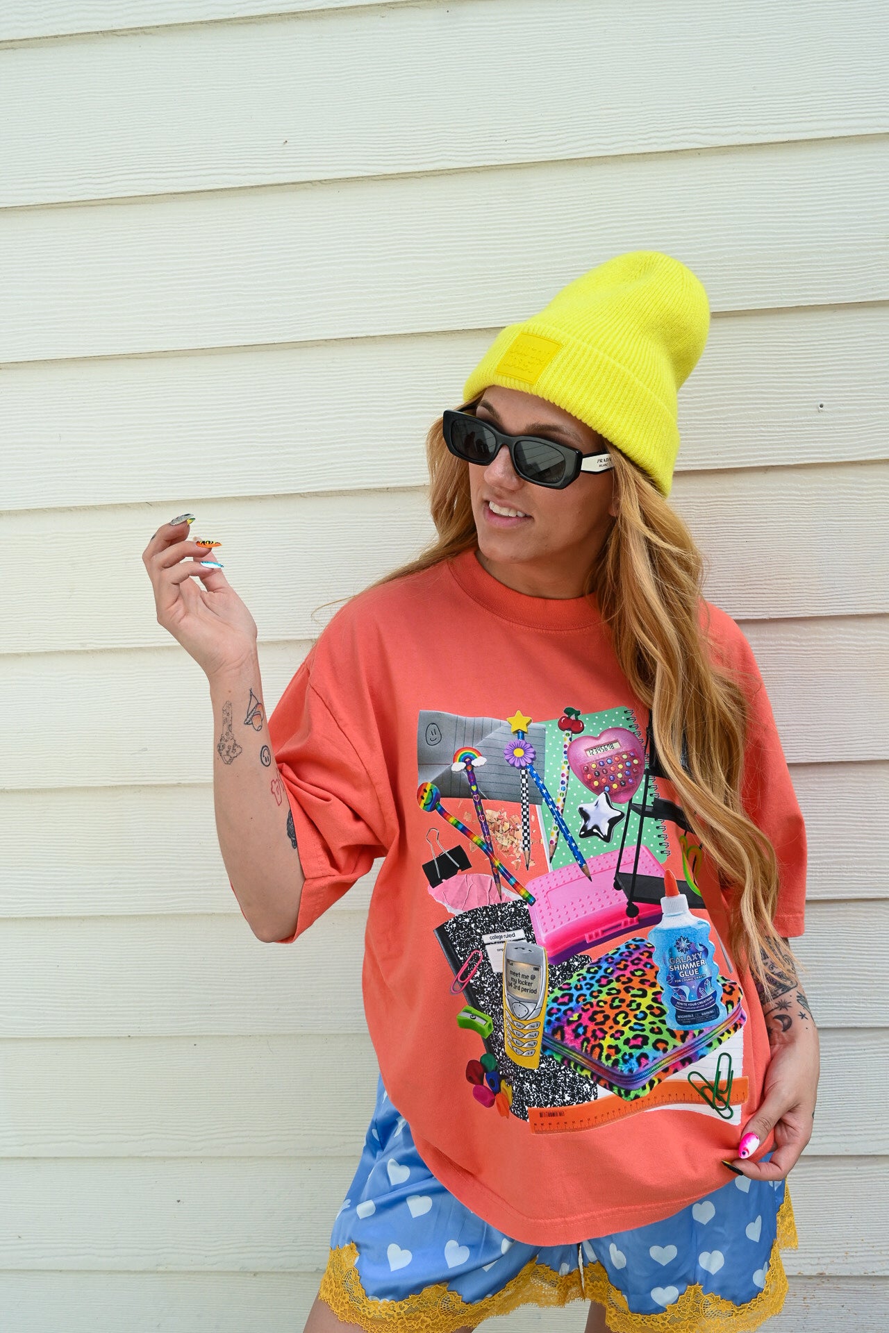 A woman wearing a bright yellow beanie, black sunglasses, a coral orange graphic t-shirt, and blue polka-dot shorts with yellow ruffle trim, standing against a white wood-paneled wall