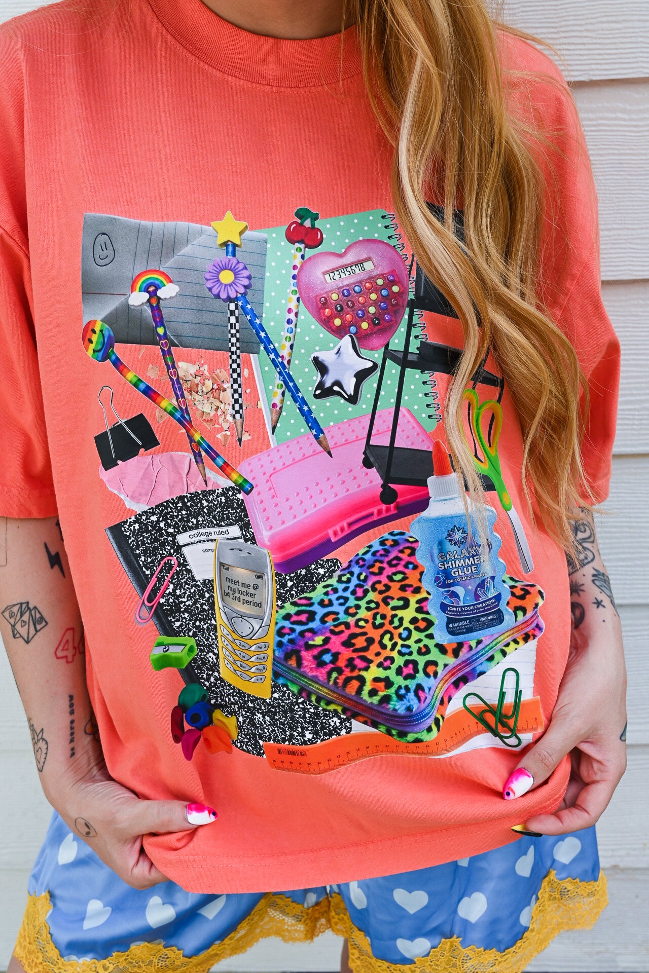 A person wearing a bright coral-orange graphic T-shirt with a colorful collage of school and stationery items, paired with blue shorts with white hearts and yellow lace trim, standing against a white wood-paneled wall