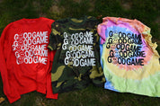 Three graphic t-shirts laid out on grass: a red sweatshirt with white text, a green camouflage t-shirt with white text, and a pastel tie-dye t-shirt with black text