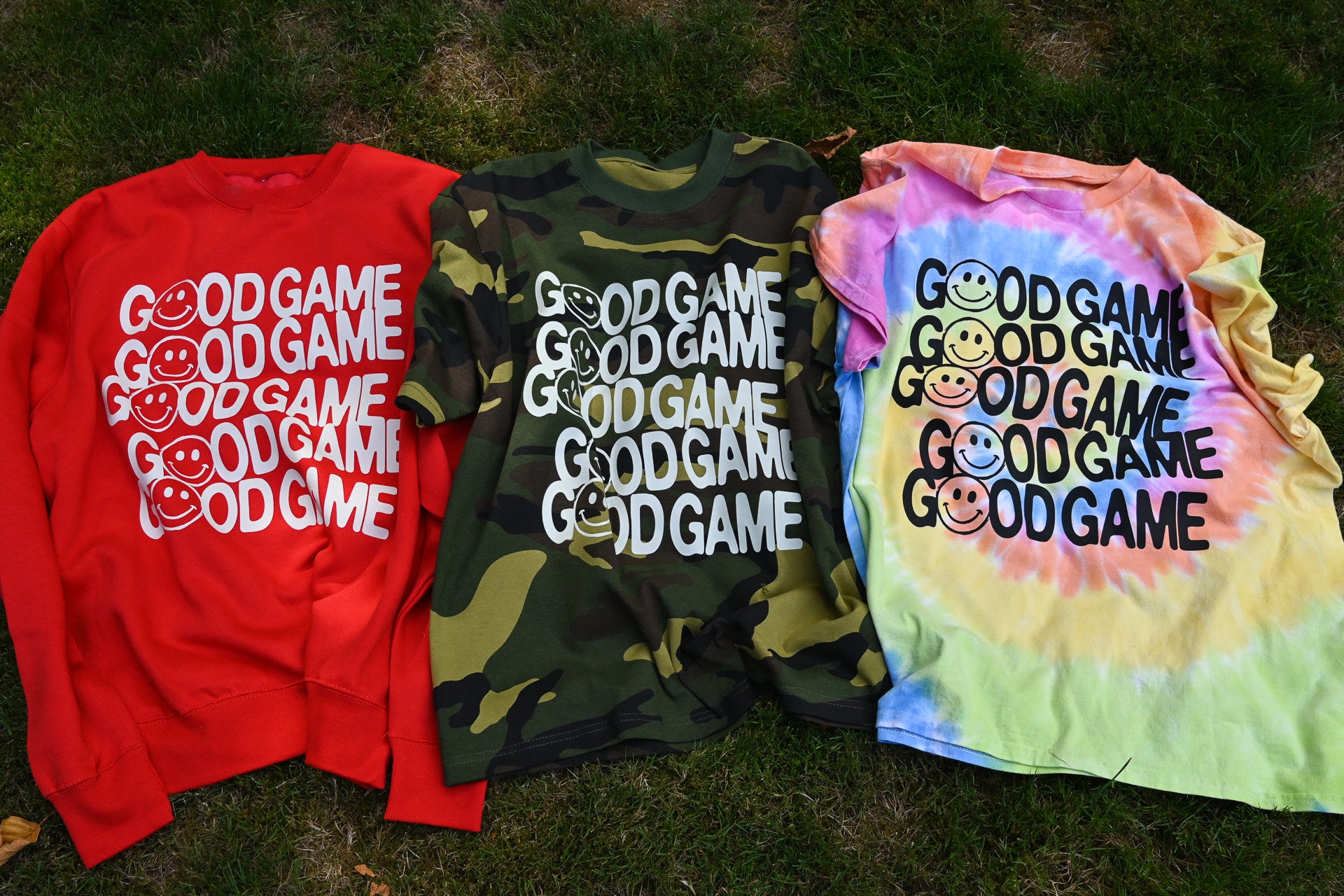 Three graphic t-shirts laid out on grass: a red sweatshirt with white text, a green camouflage t-shirt with white text, and a pastel tie-dye t-shirt with black text