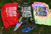 Three graphic t-shirts in red, green camouflage, and pastel tie-dye, each printed with the text 'GOOD GAME' and smiley faces, laid out on grass alongside blue sneakers