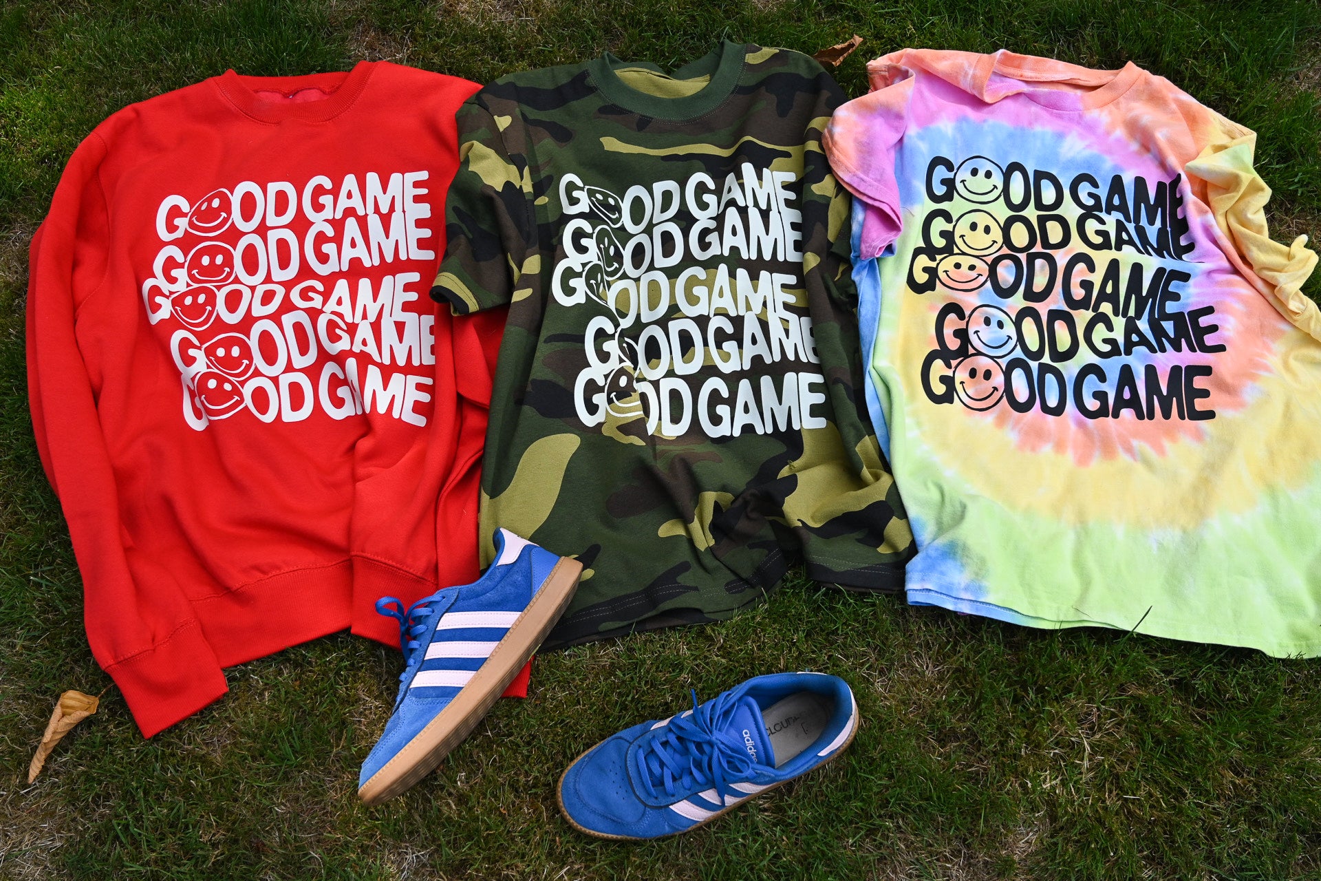 Three graphic t-shirts in red, green camouflage, and pastel tie-dye, each printed with the text 'GOOD GAME' and smiley faces, laid out on grass alongside blue sneakers