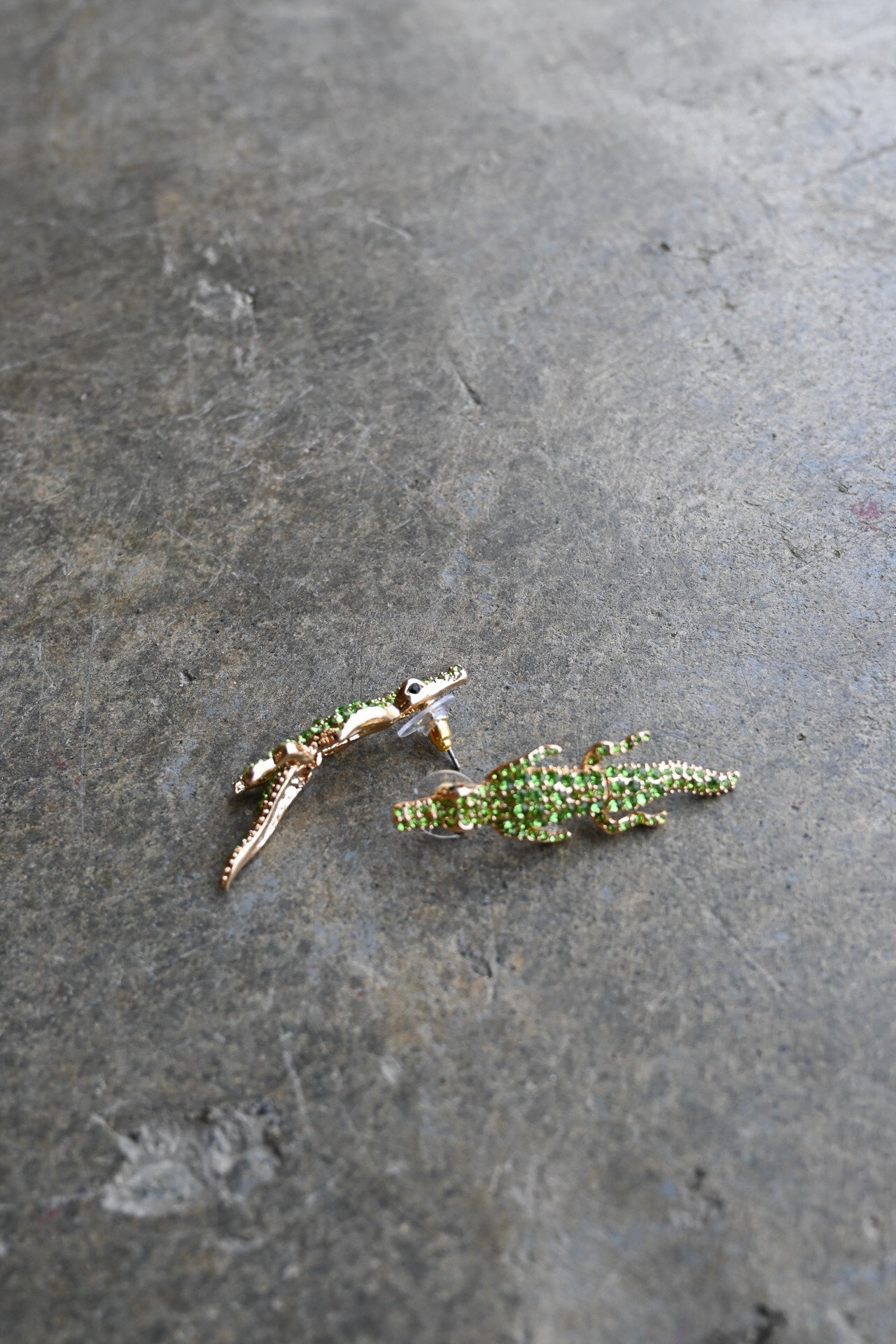 Two small lizard-shaped earrings with green textured detailing and gold-toned edges, placed on a dark gray stone surface.