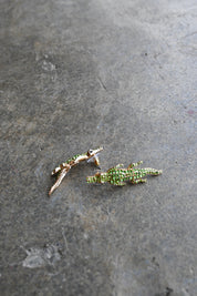 Two small lizard-shaped earrings with green textured detailing and gold-toned edges, placed on a dark gray stone surface.