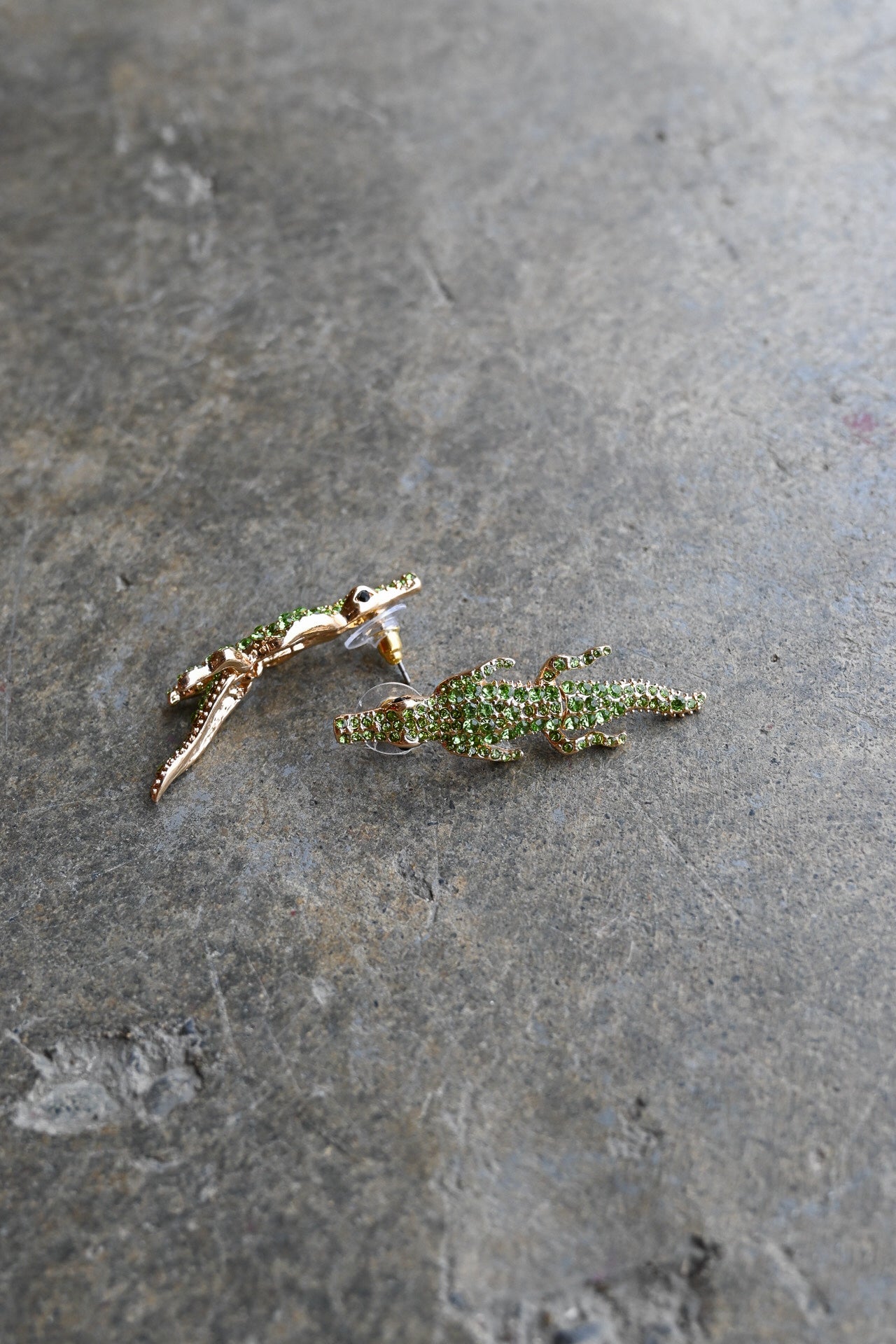 A pair of gold-toned alligator stud earrings embellished with green crystals, resting on a textured gray stone surface.