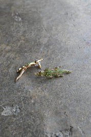 A pair of gold-toned alligator stud earrings embellished with green crystals, resting on a textured gray stone surface.