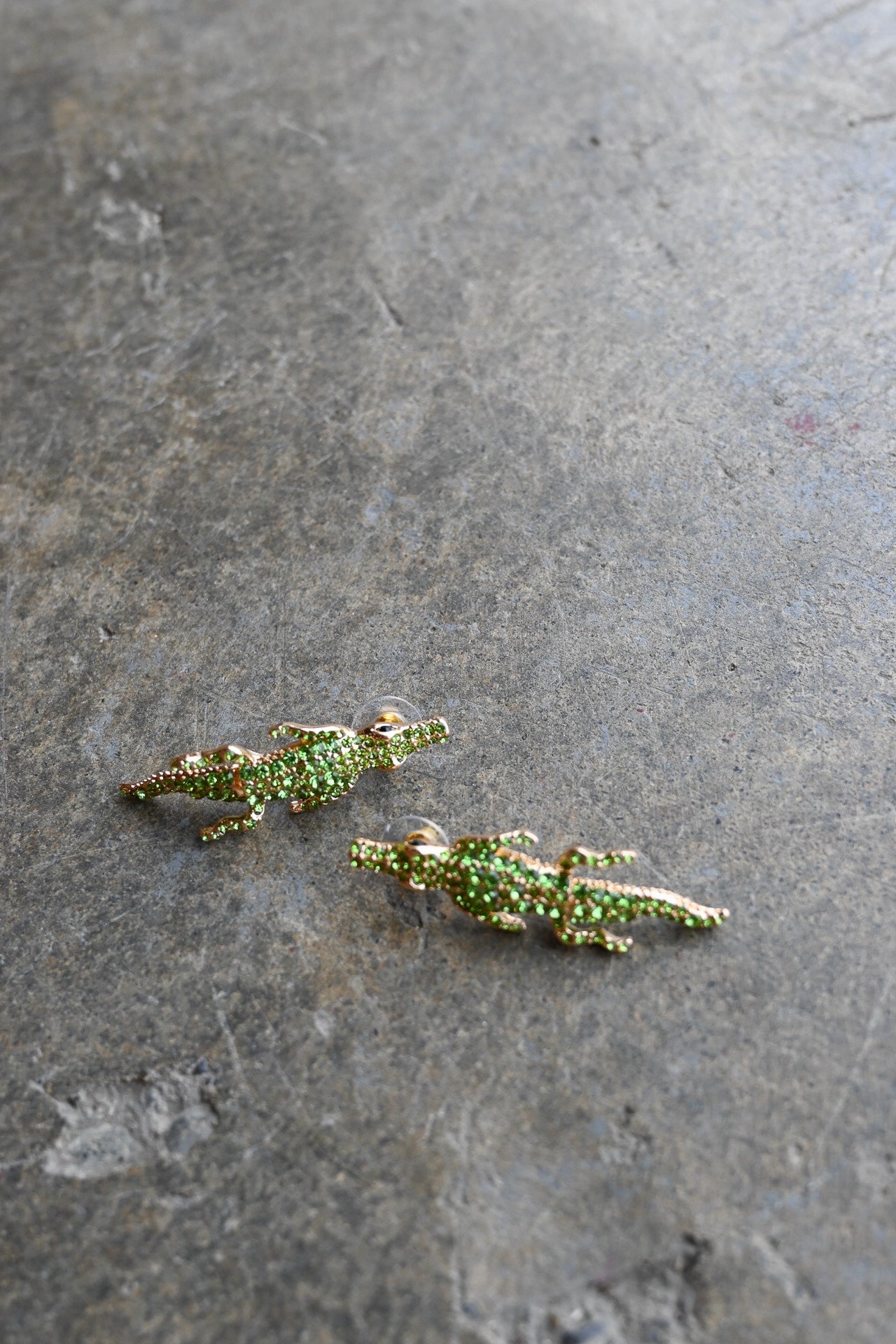 A pair of small alligator-shaped earrings covered in green rhinestones with gold-toned edges, resting on a textured gray stone surface.