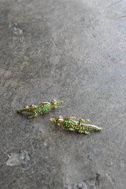A pair of small alligator-shaped earrings covered in green rhinestones with gold-toned edges, resting on a textured gray stone surface.