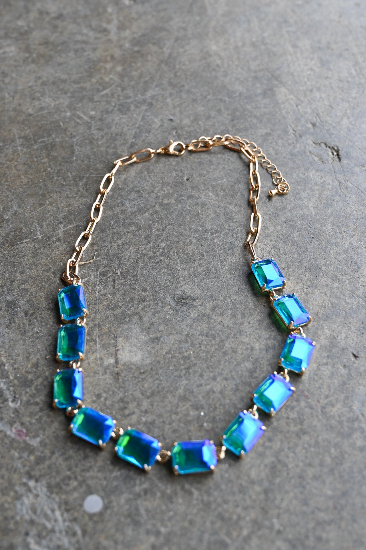 A gold chain necklace featuring a row of rectangular iridescent blue and green rhinestones, displayed on a textured gray stone surface.
