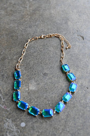 A gold chain necklace featuring a row of rectangular iridescent blue and green rhinestones, displayed on a textured gray stone surface.