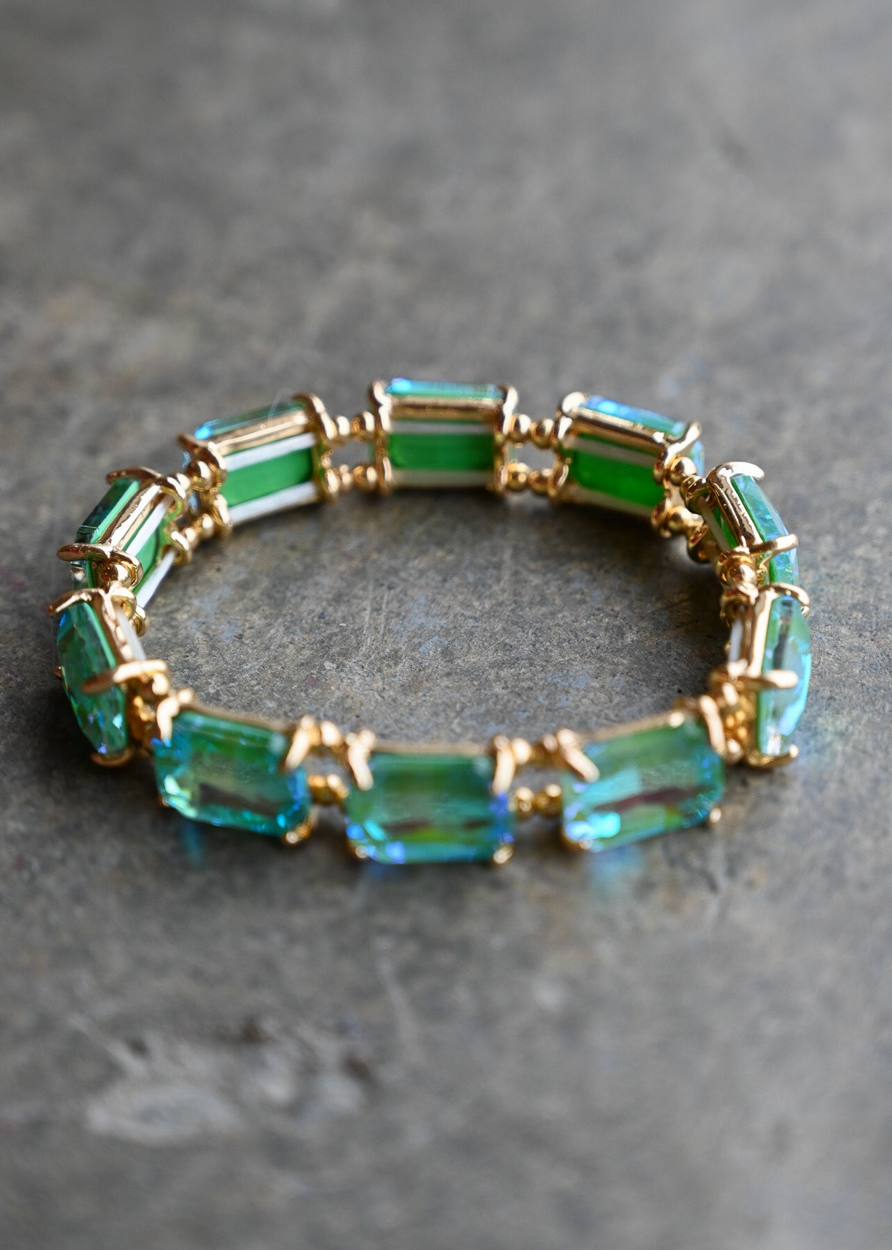 A gold-plated stretch bracelet featuring rectangular emerald-cut green and blue-green gemstones, displayed on a textured gray surface