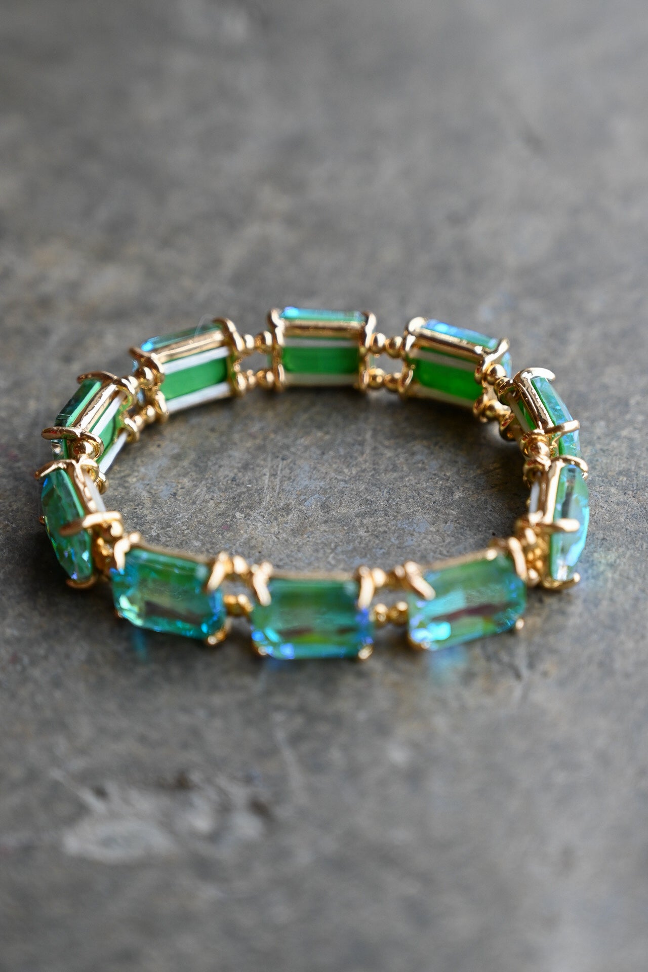 A gold-plated stretch bracelet featuring rectangular emerald-cut green and blue-green gemstones, displayed on a textured gray surface