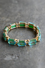 A gold-plated stretch bracelet featuring rectangular emerald-cut green and blue-green gemstones, displayed on a textured gray surface
