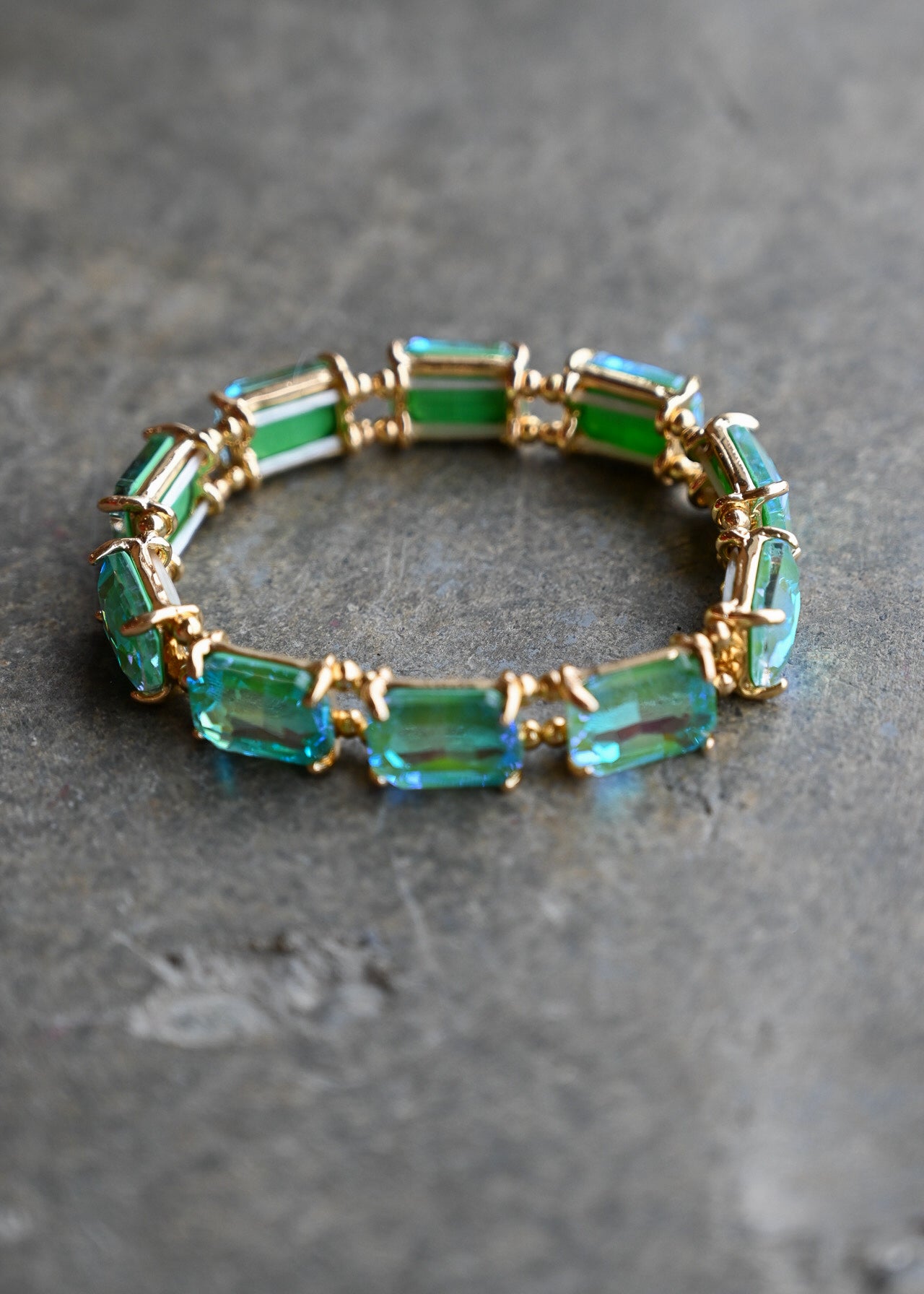A gold-plated stretch bracelet featuring rectangular emerald-cut green and blue-green crystals, displayed on a textured gray stone surface