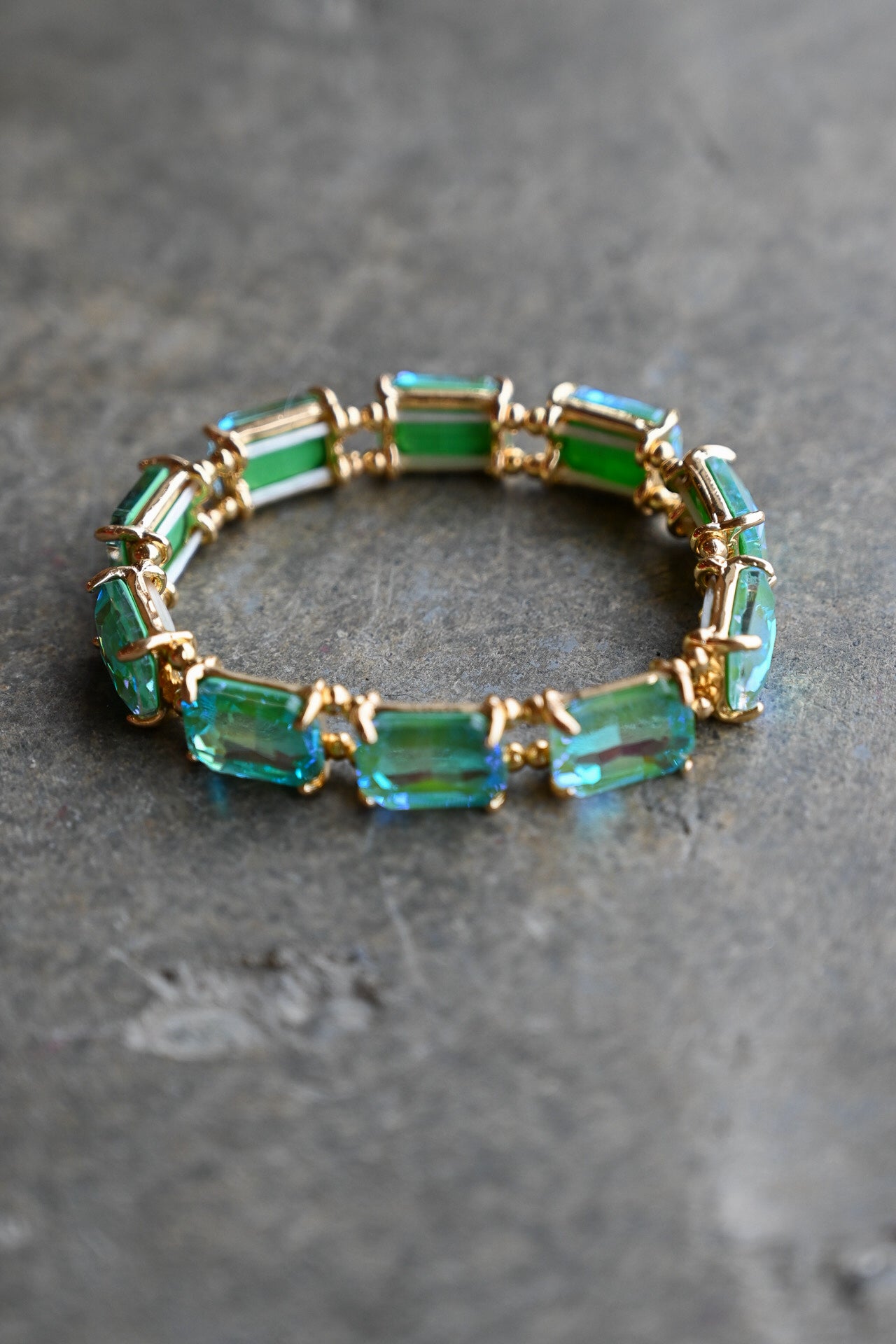 A gold-plated stretch bracelet featuring rectangular emerald-cut green and blue-green crystals, displayed on a textured gray stone surface
