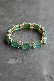 A gold-plated stretch bracelet featuring rectangular emerald-cut green and blue-green crystals, displayed on a textured gray stone surface