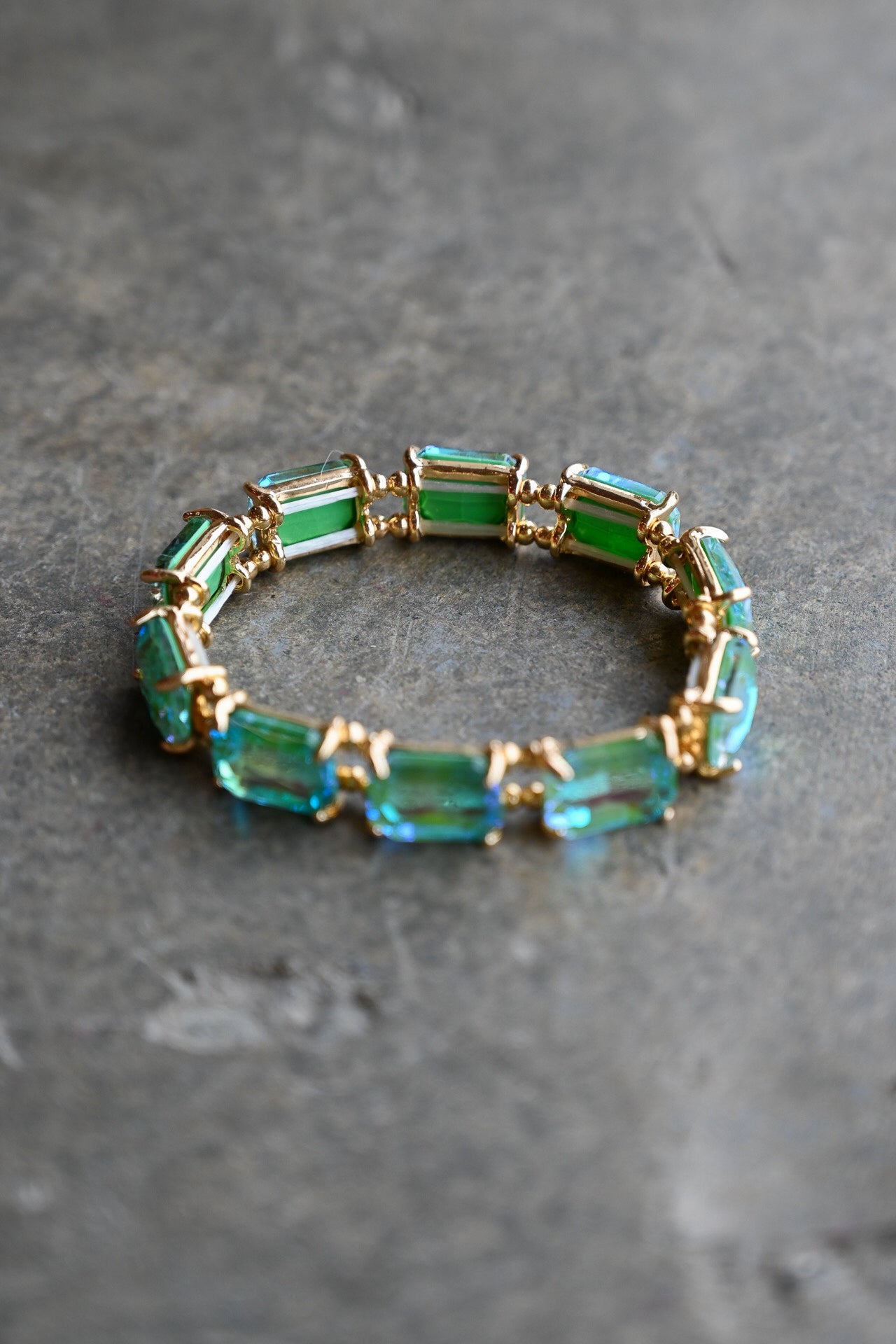 A gold-tone bracelet featuring rectangular green and blue-green gemstones, displayed on a textured gray surface
