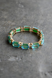 A gold-tone bracelet featuring rectangular green and blue-green gemstones, displayed on a textured gray surface