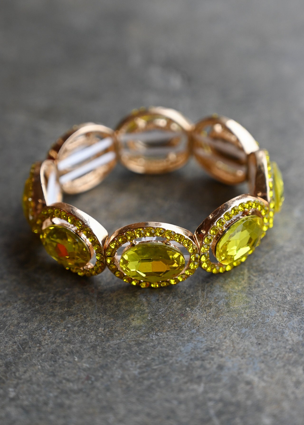 A rose gold eternity-style bracelet featuring large oval yellow gemstones surrounded by small sparkling accents, photographed on a dark textured surface