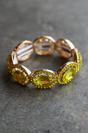 A rose gold eternity-style bracelet featuring large oval yellow gemstones surrounded by small sparkling accents, photographed on a dark textured surface