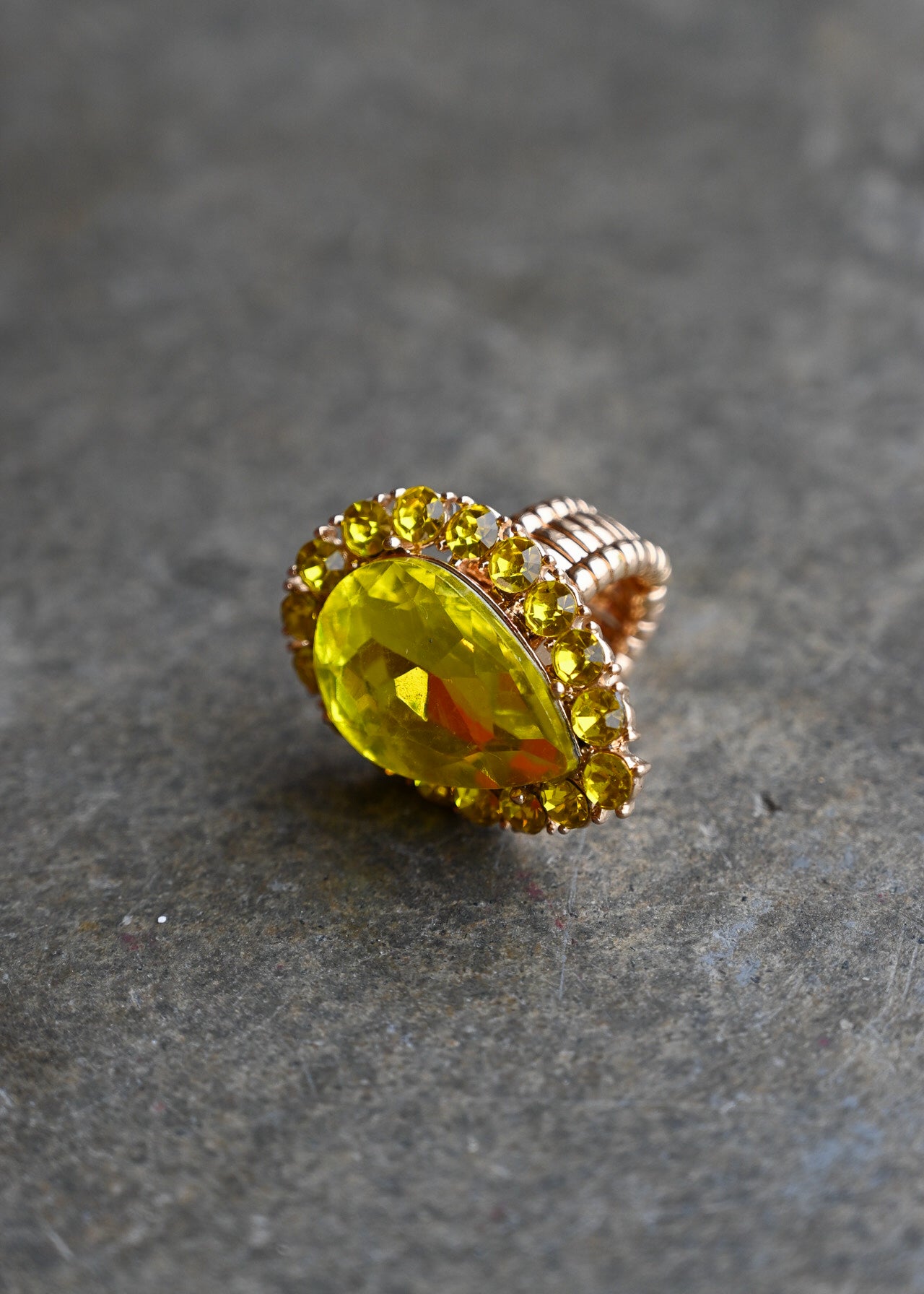 A gold ring featuring a large, faceted yellow marquise-cut gemstone surrounded by a halo of smaller yellow stones, set on a textured gray stone background