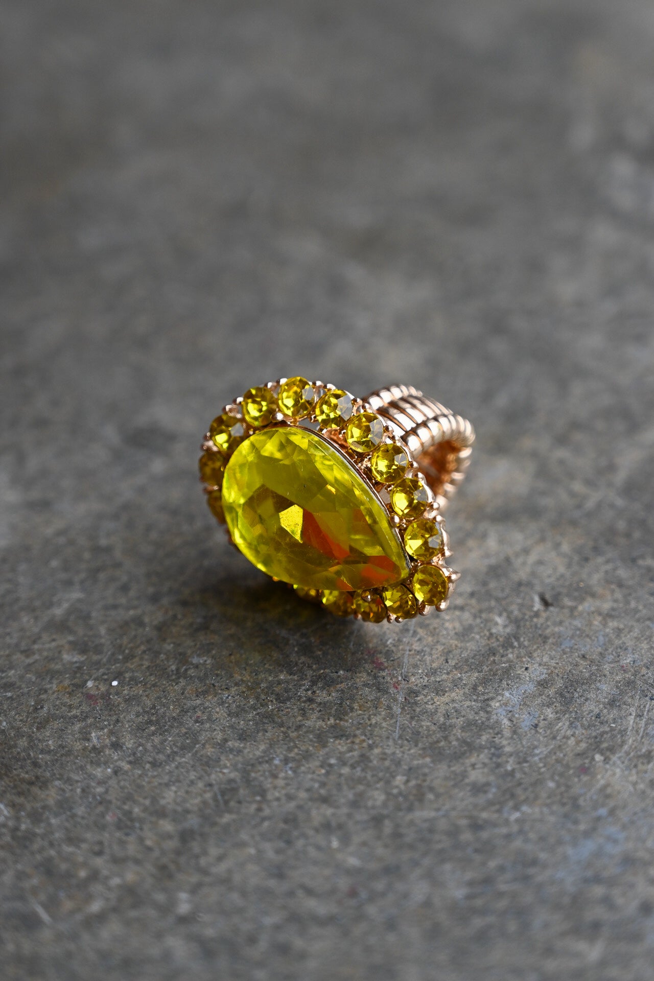 A gold ring featuring a large, faceted yellow marquise-cut gemstone surrounded by a halo of smaller yellow stones, set on a textured gray stone background