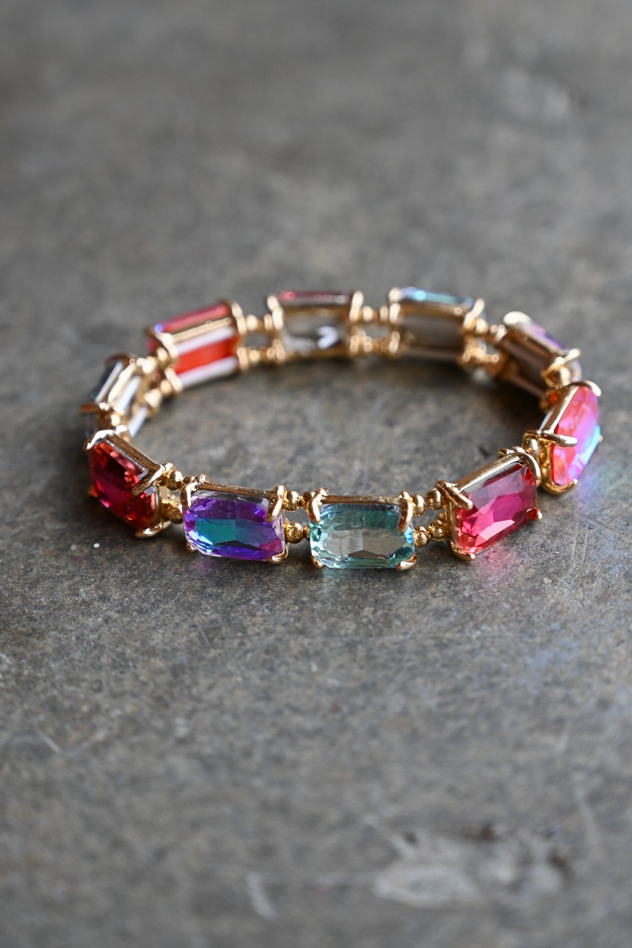 A gold-plated stretch bracelet featuring rectangular iridescent gemstones in red, blue, purple, and green, displayed on a textured gray surface