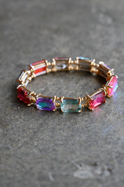 A gold-plated stretch bracelet featuring rectangular iridescent gemstones in red, blue, purple, and green, displayed on a textured gray surface