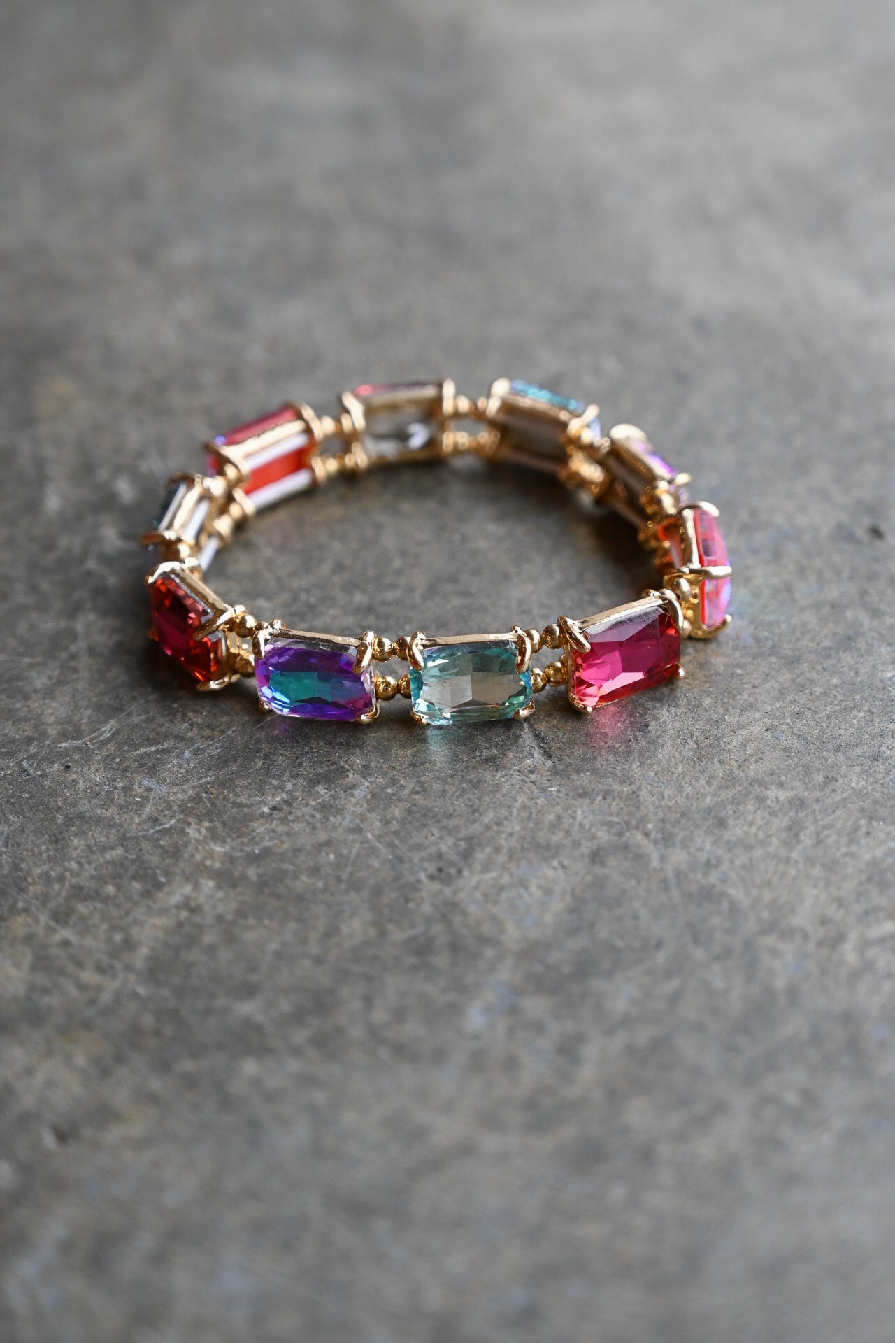 A gold-plated stretch bracelet featuring a row of colorful rectangular gemstones in red, blue, purple, and pink, resting on a textured gray stone surface