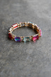 A gold-plated stretch bracelet featuring a row of colorful rectangular gemstones in red, blue, purple, and pink, resting on a textured gray stone surface
