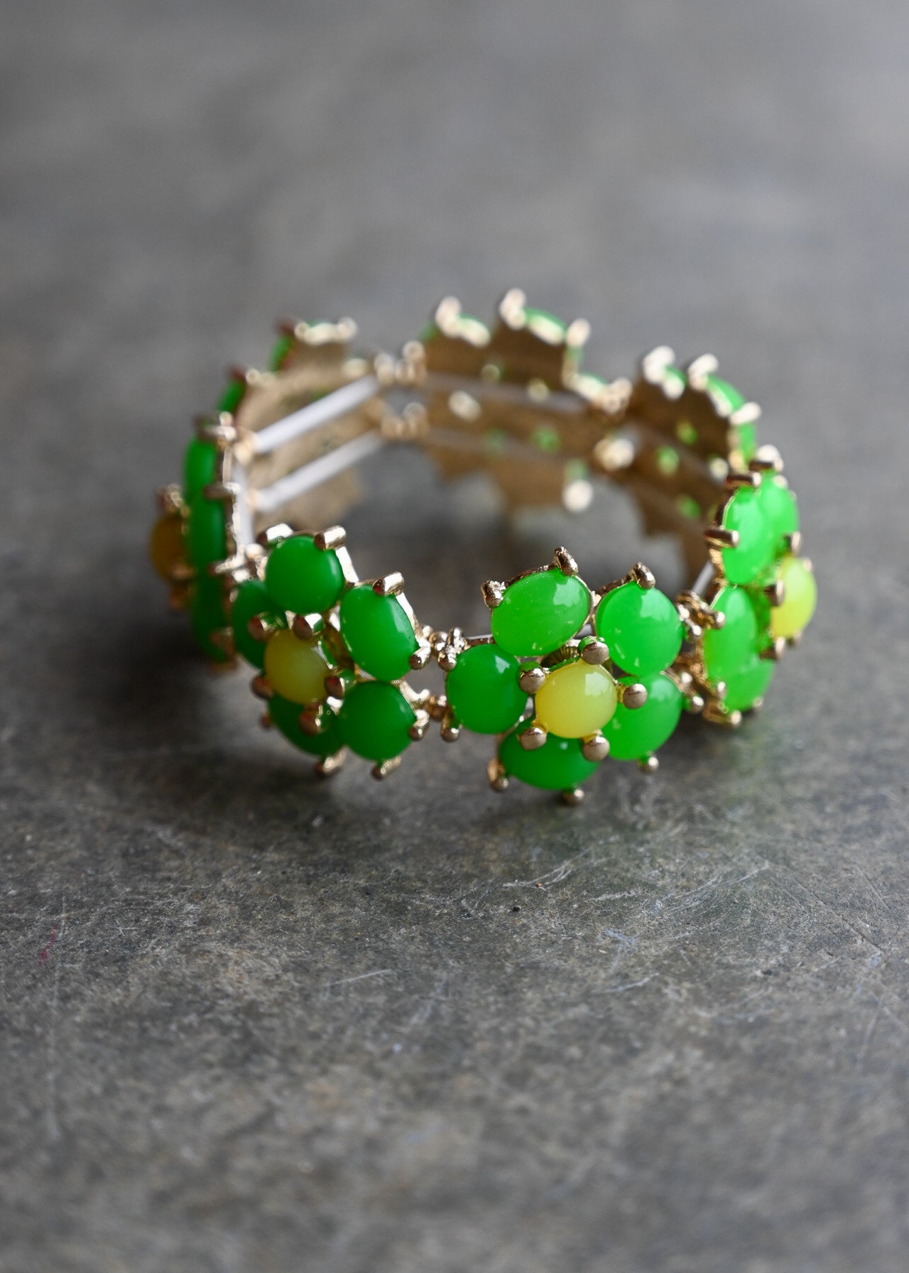 A gold-plated stretch band bracelet decorated with green and yellow gemstone clusters, resting on a textured gray surface