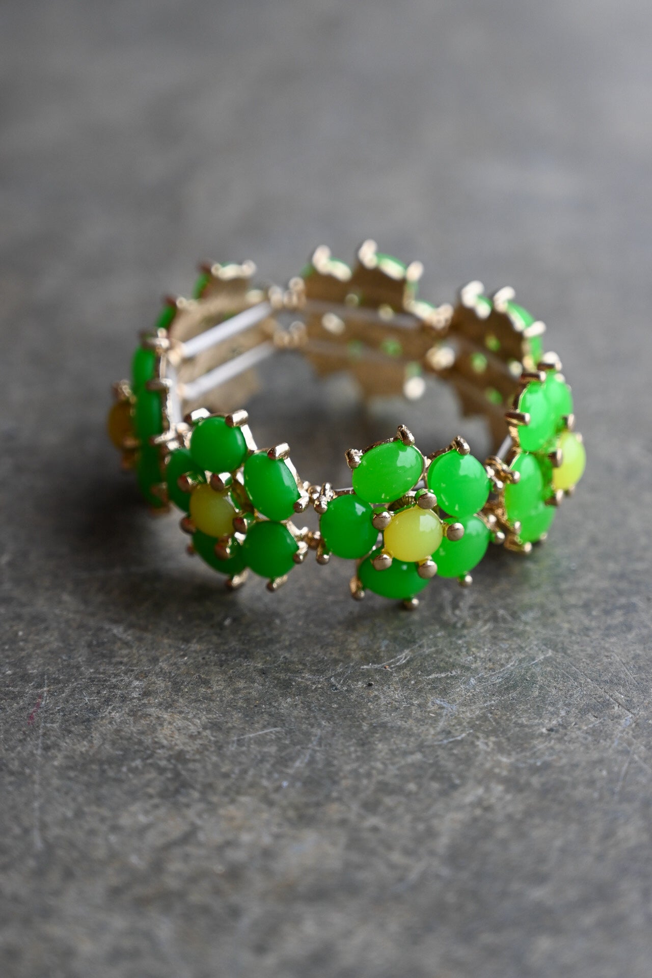 A gold-plated stretch band bracelet decorated with green and yellow gemstone clusters, resting on a textured gray surface