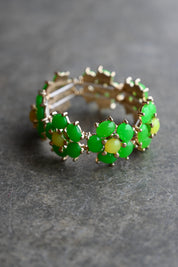 A gold-plated stretch band bracelet decorated with green and yellow gemstone clusters, resting on a textured gray surface
