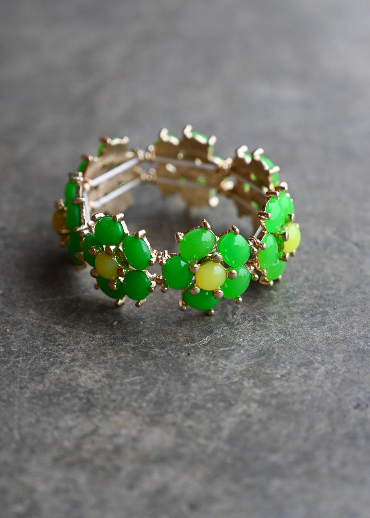 A gold-tone stretch band bracelet featuring a repeating pattern of oval green stones surrounded by small pale yellow flower centers, set against a dark gray textured background