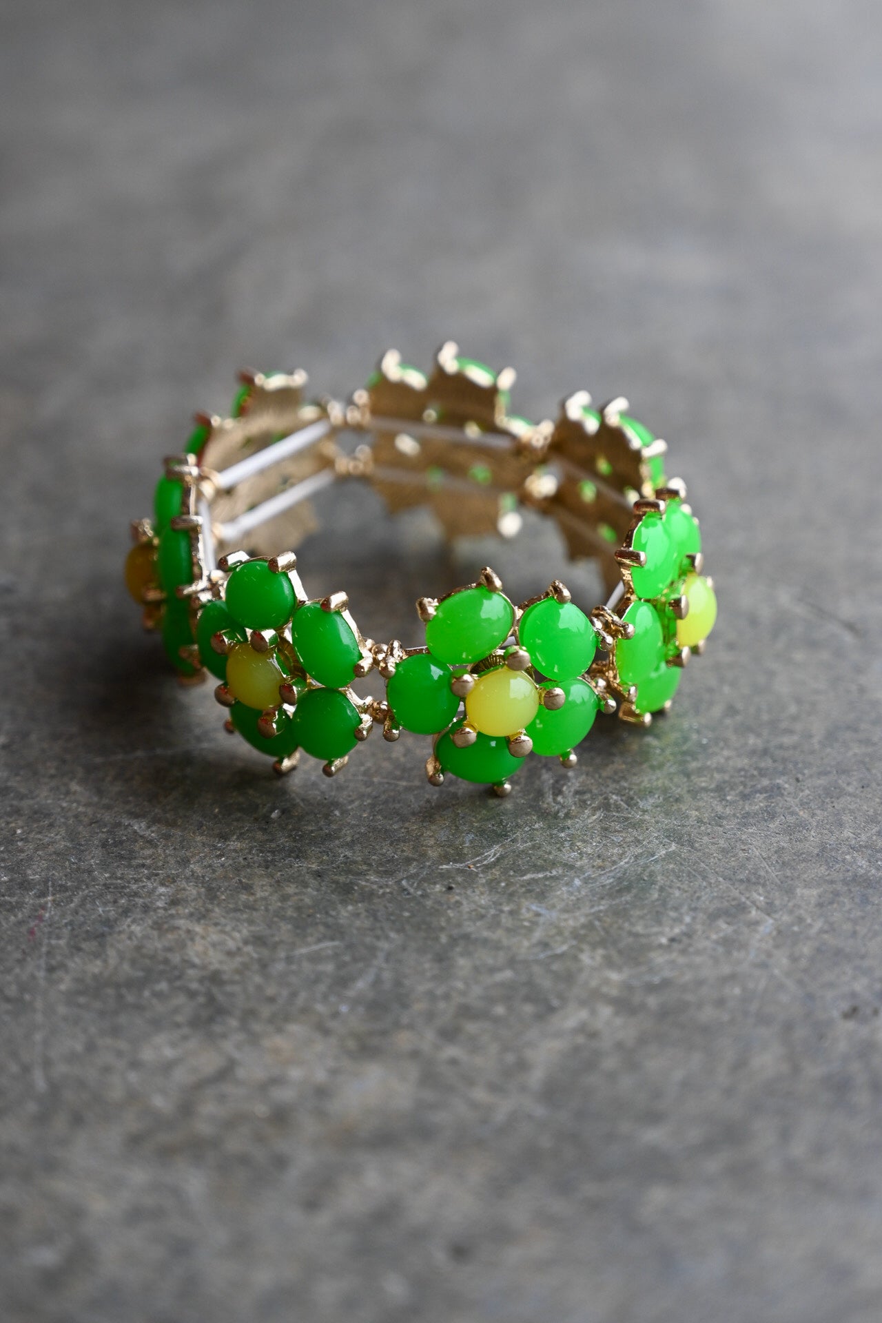 A gold-tone stretch band bracelet featuring a repeating pattern of oval green stones surrounded by small pale yellow flower centers, set against a dark gray textured background