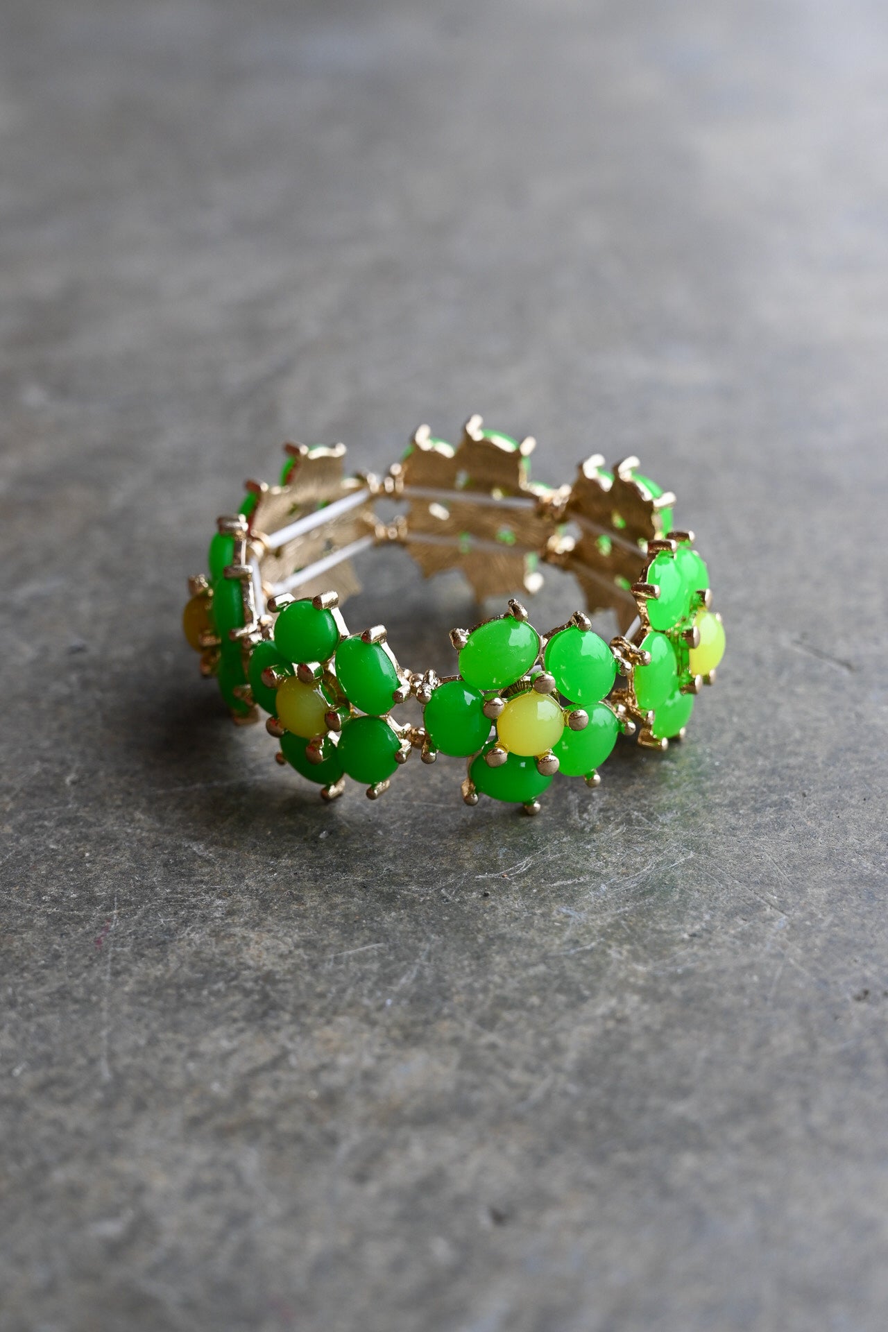 A gold-plated stretch bracelet featuring a series of green cabochon stones set in gold, with some stones showing a yellow-green iridescent shift, displayed on a textured gray surface