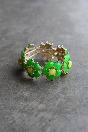 A gold-plated stretch bracelet featuring a series of green cabochon stones set in gold, with some stones showing a yellow-green iridescent shift, displayed on a textured gray surface